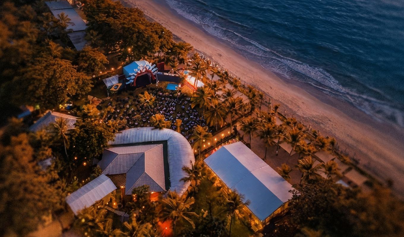 Vista aérea da Beat Beach à beira-mar, com palco, tendas e coqueiros